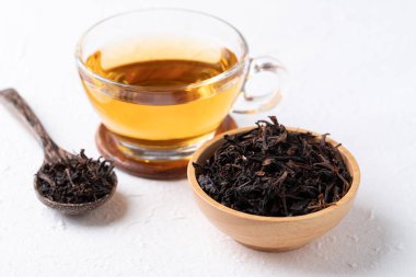 Tahta kasede kurumuş siyah çay yaprakları ve beyaz arka planda bardakta sıcak çay, sağlıklı bitkisel içecekler.