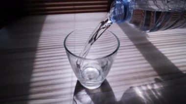 Plastik şişeden akan suyun yavaş çekimde pencerenin yakınındaki ahşap bir masanın camına boşaltılması, güneş ışığının içeri akması, yukarı eğilmesi ve İzleme Çekimi