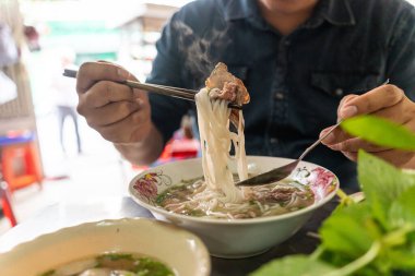 Yemek çubuğu ve kaşık kullanan bir adam geleneksel Pho Bo Vietnam çorbası yiyor. Metal bir masada sığır eti ve pirinç eriştesi, yerel bir restoranda gerçek sahne, seçici bir odak noktası.