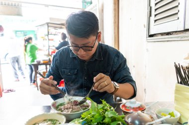 Yemek çubuğu ve kaşık kullanan bir adam geleneksel Pho Bo Vietnam çorbası yiyor. Metal bir masada sığır eti ve pirinç eriştesi, yerel bir restoranda gerçek sahne, seçici bir odak noktası.