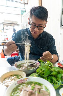 Yemek çubuğu ve kaşık kullanan bir adam geleneksel Pho Bo Vietnam çorbası yiyor. Metal bir masada sığır eti ve pirinç eriştesi, yerel bir restoranda gerçek sahne, seçici bir odak noktası.