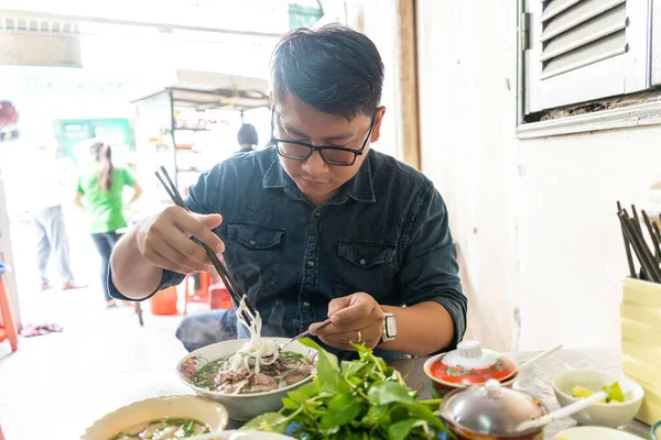 Yemek çubuğu ve kaşık kullanan bir adam geleneksel Pho Bo Vietnam çorbası yiyor. Metal bir masada sığır eti ve pirinç eriştesi, yerel bir restoranda gerçek sahne, seçici bir odak noktası.