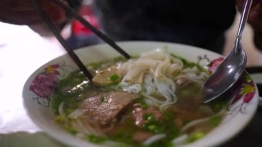 Yemek çubuğu ve kaşık kullanan bir adam geleneksel Pho Bo Vietnam çorbası yiyor. Metal bir masada sığır eti ve pirinç eriştesi, yerel bir restoranda gerçek sahne, seçici bir odak noktası.