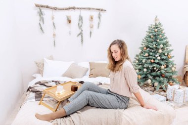 Orta yaşlı modern bir kadın İskandinav tarzında Noel 'de yatakta oturuyor. Kız gülümsüyor, tarafsız renk süveter giyiyor, gri pantolon giyiyor. Beyaz bir duvarda, çelenkle süslenmiş ahşap bir engel, Noel ağacı