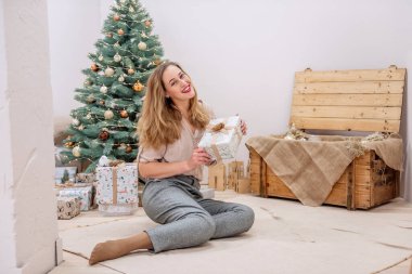 Modern çağdan kalma şık bir kadın yapay Noel ağacında oturuyor, elinde bir hediye tutuyor, gülümsüyor. Dudaklarındaki kırmızı ruj. Neşeli bir çevre dostu iç mekan. Boşluğu kopyala, potrtait 'i kapat