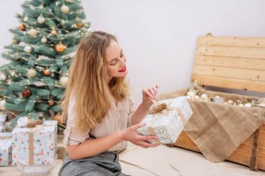 Modern çağdan kalma şık bir kadın yapay Noel ağacında oturuyor, elinde bir hediye tutuyor, gülümsüyor. Dudaklarındaki kırmızı ruj. Neşeli bir çevre dostu iç mekan. Boşluğu kopyala, potrtait 'i kapat