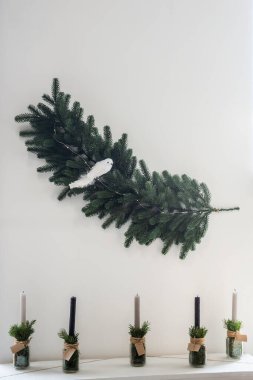 Şöminenin yanında minimalist Noel süslemesi. İskandinav kırsal tarzı. Kuşlu çelenk beyaz duvarda asılı, el yapımı, siyah beyaz mumlu cam şamdanlar.