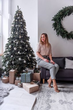 Kırmızı dudaklı, narin gömlekli ve gri pantolonlu şık bir milenyum kadını Noel ağacının yanındaki gri renkli koltukta oturuyor. İskandinav iç mimarisi. Moda Yeni Yıl