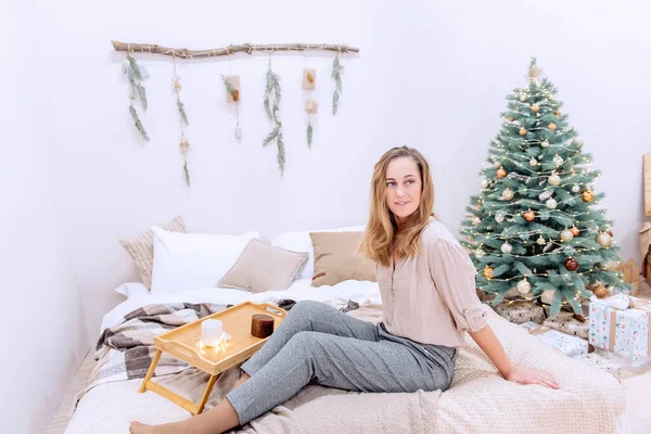 現代の中年女性のベッドの上にスキャンディナヴィアスタイルのクリスマスに座っている 女の子は 中性色のセーター グレーのズボンを着て笑っている 白い壁には ガーランド クリスマスツリーで飾られた木製のスナック — ストック写真