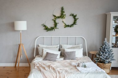 Beyaz dövmeli demir yatak. Sütlü atımlar ve Noel ağacının yanındaki ahşap zeminde yastıklar. Gri beton duvarında çelenk asılı doğal yeşil çelenkler. Moda, modern iç mimari