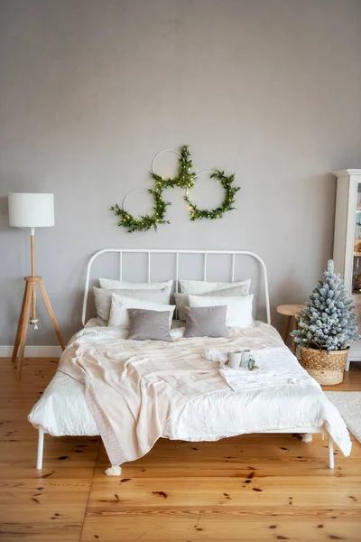 White wrought iron bed with textured milky throws and pillows on a wood floor next to a Christmas tree. Natural green wreaths with a garland hang on a gray concrete wall. Trendy, modern interior