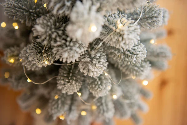 一株白雪覆盖的人造圣诞树的顶部 装饰着明亮的花环 矗立在木制的层压板地板上 度假卡 简约主义风格 模糊的纹理背景 复制空间 — 图库照片