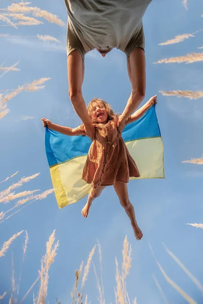 Man throws up little girl who holds blue yellow flag of Ukraine in hands against background clear sky. The long-awaited meeting of father and daughter among spikelets. Come back alive, fatherhood