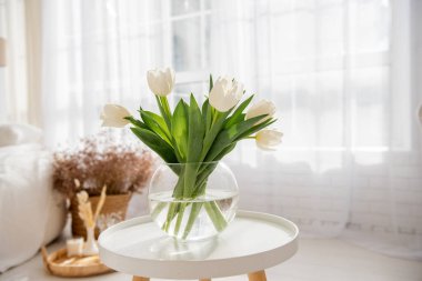 On round wooden table stands glass vase with a bouquet of white tulips. Airy, light interior in bright colors. Freshness, purity of natural colors. Spring time. Flowers for a holiday, mood. Copy space