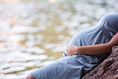 Close-up portrait of the belly of middle aged pregnant woman in blue dress, sits on rock by the sea. Millennial girl travels in pregnancy on vacation. Family holiday on the ocean. Natural texture.