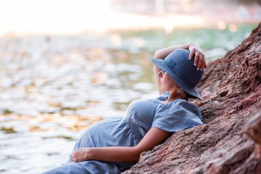 Middle aged pregnant woman in blue dress, hat sits on rock by the sea. Millennial girl travels in pregnancy on vacation. Family holiday on the ocean. Natural texture. Merging with nature. Copy space