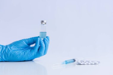 On white isolated background, hands of doctor in medical blue gloves hold ampoule vaccine against virus coronovirus. Syringe with antidote, pain pills. Fighting pandemic, epidemic. Mock up, copy space