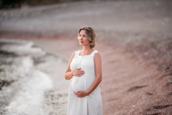 Portrait of middle aged pregnant woman in white sundress. Expectant mother walks along the seashore at sunset. Concept of travel in pregnancy, healthy vacation. Enjoyment of life. Purity, copy space