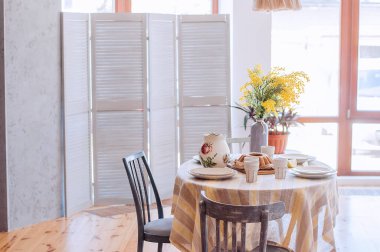Spring, Easter interior in Scandinavian style. Rustic Living room with a bright wardrobe, yellow mimosa in vase. The table is festively set with ceramic dishes, with a cake on a tray. macrame details