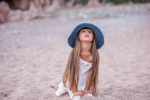 青い帽子の少女は 白いドレスは砂の上で再生され 海岸沿いの小さな小石 子供との休日 子供は海のそばに石の塔を立っている 夏の子供時代 スペースのコピー — ストック写真