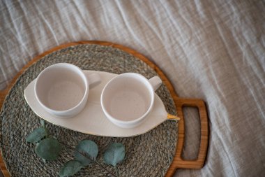 On the Linen bedspread lies a wooden round tray, on it are two empty ceramic cups, green eucalyptus. Stylish, trendy morning. Textural, Scandinavian Rustic interior. Light gray background. Copy space