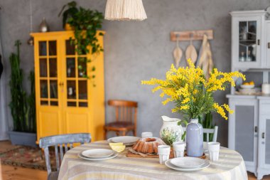 Spring, Easter interior in Scandinavian style. Rustic Living room with a bright wardrobe, yellow mimosa in vase. The table is festively set with ceramic dishes, with a cake on a tray. macrame details