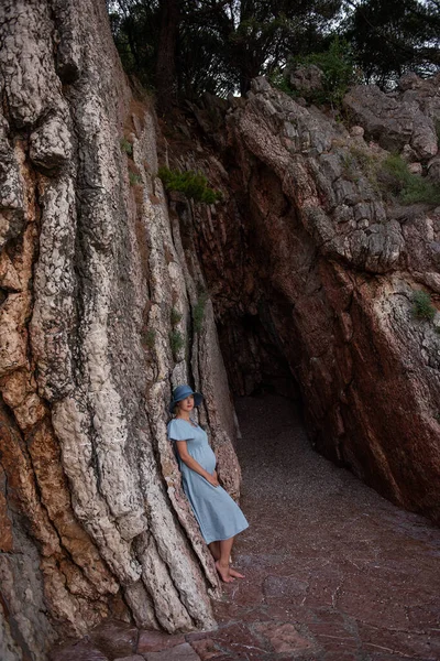 Pregnant Millennial Woman Blue Dress Stands Rocks Seashore Hugging Belly — Stockfoto