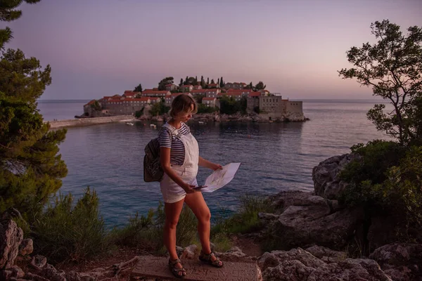 Middle aged pregnant woman in vest holds map, phone in hands. Traveler with backpack looks at the island of Sveti Stefan at sunset in Montenegro. Travel motherhood pregnancy concept. Summer vibes