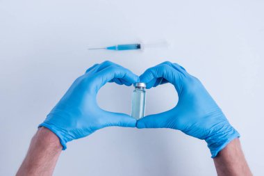 On white isolated background, the doctors hands in medical blue gloves depict heart shape gesture. Doctor is holding an ampoule with vaccine against virus. Blurred syringe with medicine. Copy space