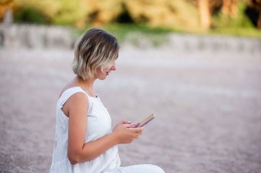 ?lose-up portrait of a woman in a white sundress sits on the sand by the sea, typing a message on a smartphone. Green phone screen mockup. Internet connection in tourism. Faceless, copy space, mock up