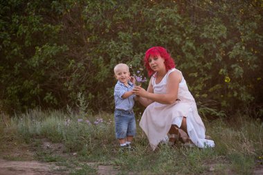 Küçük çocuk annesine sevgiyle bir buket yabani çiçek verdi. Anneler Günü konsepti. Pembe saçlı, çocuklu genç bir kadın parkta yürüyor. Botanik doğa geçmişi. Boşluğu kopyala