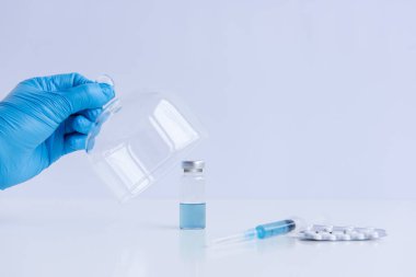 On white isolated background stands the vaccine with syringe, pills. Doctors hand in blue medical glove opens programmed protective dome. Cure against viruses, treatment of people. Mock up, copy space