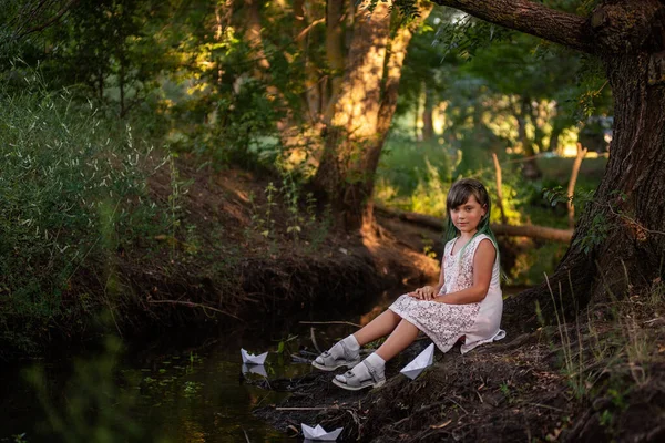 ヴィンテージのドレスで緑の髪の少女の肖像画は 手に白い折り紙の紙のボートを保持し 川の銀行に座っている 子供は森の中で船を元気に起動します 旅行のコンセプト — ストック写真