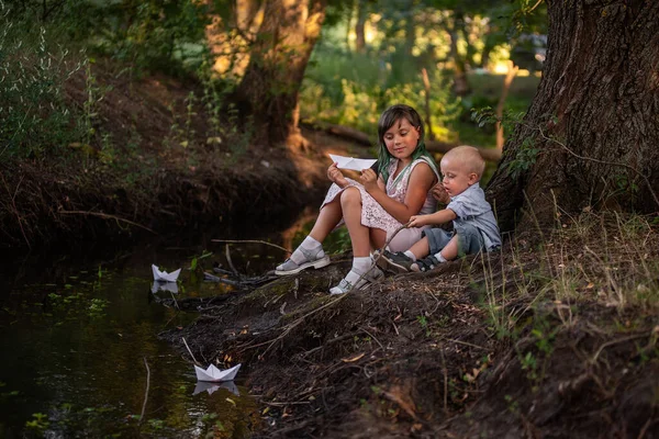Dziewczyna Chłopiec Siedzą Brzegu Rzeki Uruchamiając Białą Papierową Łódkę Origami — Zdjęcie stockowe