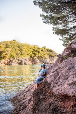 Middle aged pregnant woman in blue dress, hat sits on rock by the sea. Millennial girl travels in pregnancy on vacation. Family holiday on the ocean. Natural texture. Merging with nature. Copy space