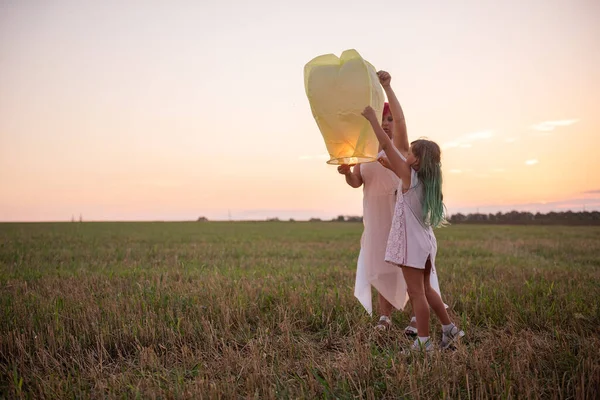 Diversity woman and girl light a yellow paper lantern in the sky at sunset in a field. Relationship between mother and daughter. Teamwork. Making wishes. Concept of hope, faith, travel. Copy space