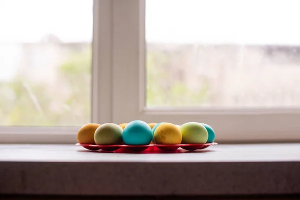 On a red, round stand near the window, bright Easter eggs lie on a marble table. Homogeneous lifestyle background. Easter traditions to paint eggs. Beautiful light shadow.