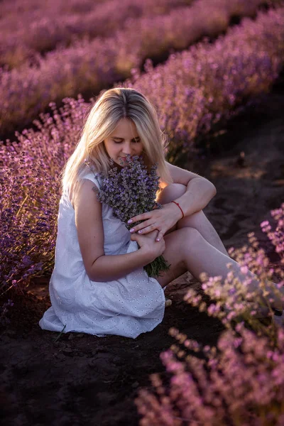 白いドレスを着た若い女性が畑に紫色のラベンダーの列に座っています 千年紀の少女は日没時に花束を手に持っている アレルギーの概念 天然の香水の製造 田舎旅行 — ストック写真