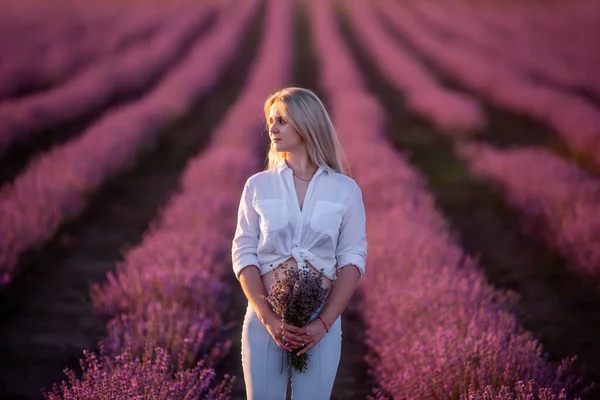 白いシャツの若い女性は 青いジーンズはフィールドに紫色のラベンダーの行に滞在します 千年紀の少女は日没時に花束を手に持っている アレルギーの概念 天然香料の製造 — ストック写真