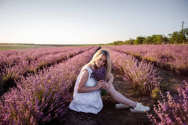 白いドレスを着た若い女性が畑に紫色のラベンダーの列に座っています 千年紀の少女は日没時に花束を手に持っている アレルギーの概念 天然の香水の製造 田舎旅行 — ストック写真