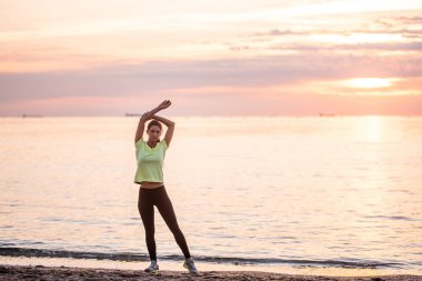 Spor giysili genç bir kadın sabahın erken saatlerinde güneş doğarken deniz kıyısında ısınıyor. Uzun saçlı ince yapılı kız esniyor. Açık hava sporları, deniz kenarında. Spor dersleri, vücut bakımı, akıl sağlığı.