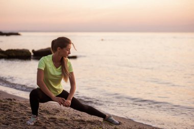 Spor giysili genç bir kadın sabahın erken saatlerinde gün doğumunda deniz kenarında mekik çeker. Uzun saçlı ince yapılı kız esniyor. Açık hava sporları, deniz kenarında. Spor dersleri, vücut bakımı, akıl sağlığı.