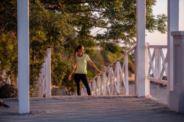 Spor kıyafetli genç bir kadın antrenmandan sonra dinleniyor şafak güneşinin altında tahta merdivenlerde yürüyor. Şehir caddesinde koşuyor. Spor ve spor konseptinde poz veren bir kız. Doğa. Boşluğu kopyala