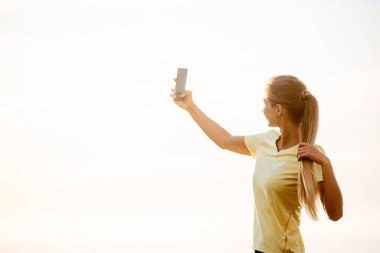 Spor kıyafetli genç bir kadının yakın plan portresi, elinde akıllı telefon, spor çalışmaları arasında portakal rengi bir gökyüzünün arka planına karşı selfie çekiyor. Spor ve iletişim kavramı. Siber uzay