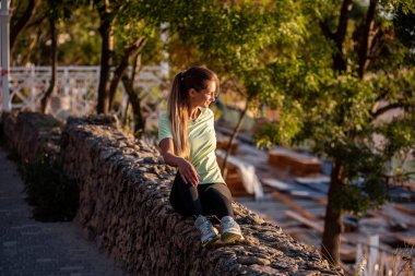 Spor sporcusu kadın gün batımında dinleniyor. Aktif bir koşu antrenmanından sonra kayaların üzerinde oturan bir kız. Açık havada çekilen mutlu sporcu kadın, deniz kenarında spor yaptıktan sonra mola veriyor.