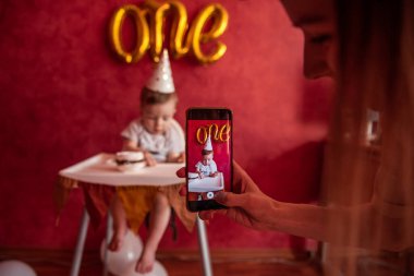 Annenin eli, oğlunun akıllı telefondaki fotoğraflarını çekiyor. Bir yaşında şapkalı, elinde mumla elinde doğum günü pastası tutan bir çocuk. Kırmızı arka planda altın folyo balonlar bir. Sosyal Medya