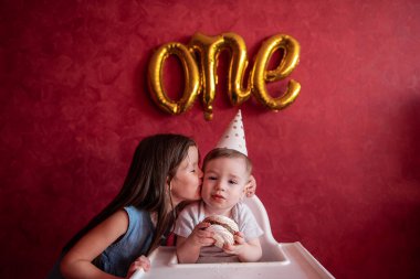 Ablası küçük erkek kardeşini ilk doğum gününde tebrik eder. Kız küçük oğlanı öpüyor. Çocuklar kırmızı arka planda folyo balonlarla eğleniyor. Pastalı ev partisi. Boşluğu kopyala