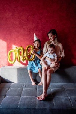 Genç anne, gri kanepedeki kırmızı duvarın yanında küçük çocuklarla birlikte kanepede oturuyor. Bir ebeveyni olan aile, bebeğin ilk yılını kutlar. Büyük ablamın elinde Folyo Balonları Bir var. Evde yaşam tarzı partisi..
