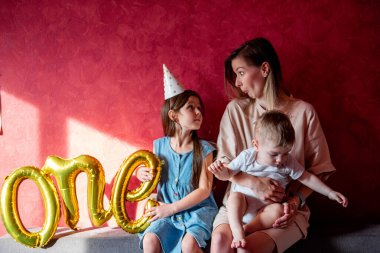 Annem, gri kanepedeki kırmızı duvarın yanında küçük çocuklarla kanepede oynaşıyor. Bir ebeveyni olan aile ilk yıl bebeklerini kutlar. Büyük ablamın elinde Folyo Balonları Bir var. Evde yaşam tarzı partisi.