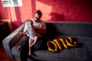 Farklılık abidesi babası, oğluyla kırmızı duvara yaslanmış gri koltukta eğleniyor. Bir adam küçük bir çocuğun ilk doğum gününü kutlar. Birinci balonların altın yazıtları. Ev hayatı partisi